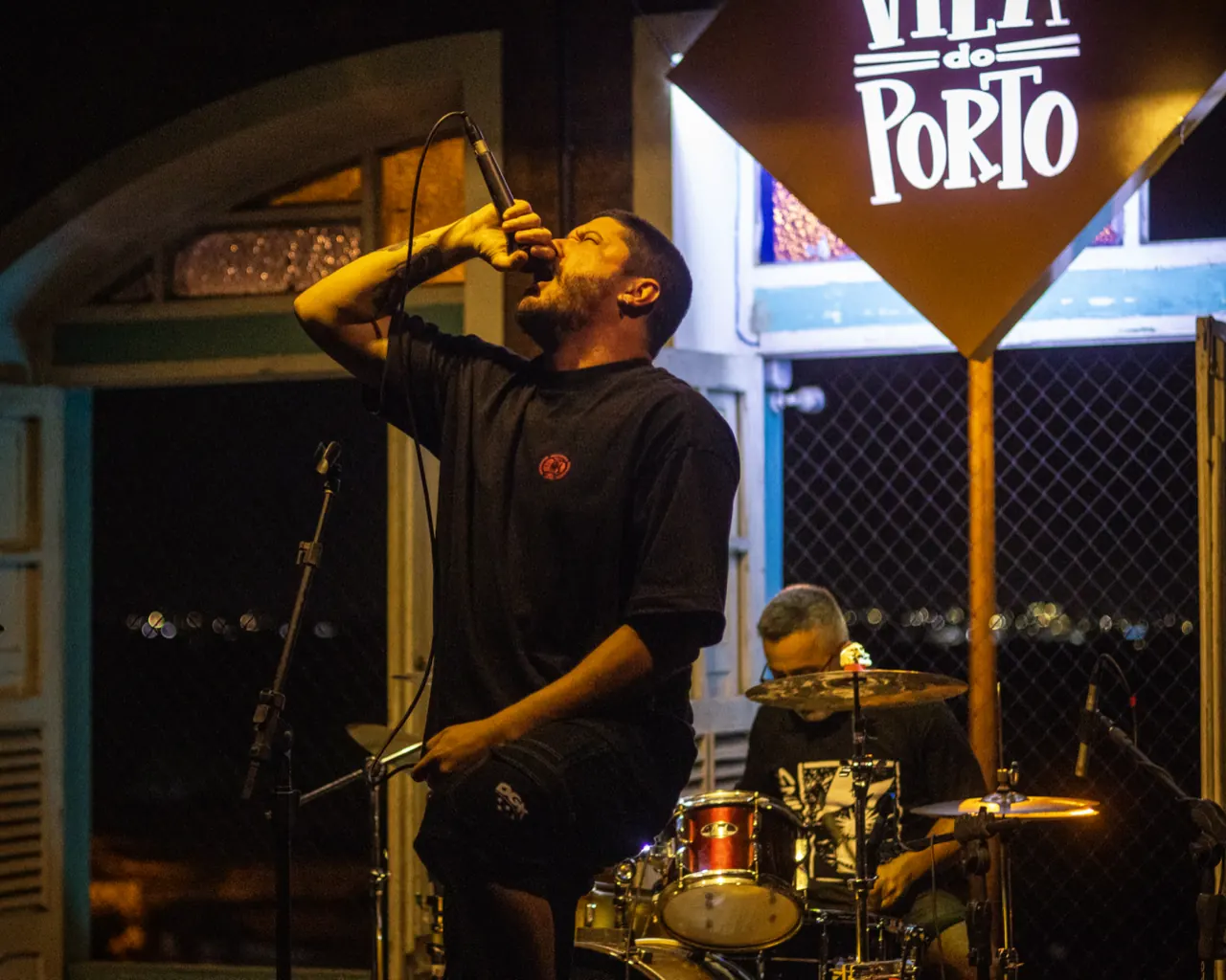 Vocalista canta com o microfone erguido, com a bateria e a placa do Vila do Porto ao fundo.